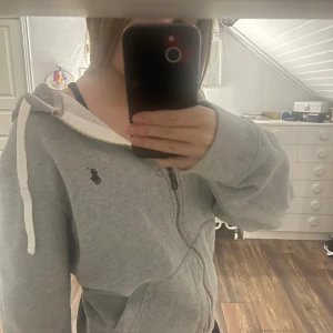 Grå hoodie från Polo Ralph Lauren - Säljer en grå hoodie från Polo Ralph Lauren med dragkedja och huva med vit snörning. Tröjan har lång ärm, fickor framtill och den klassiska Polo-loggan broderad på bröstet. Använd men inte några skador på tröjan! Säljer eftersom den är för stor🫶