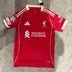 Liverpool röd matchtröja Adidas Isak 9 - Liverpool FCs officiella röda matchtröja från Adidas med vita detaljer och tryck. Kortärmad modell med klubbmärke, sponsorlogga och Champions League-märke på ärmen. Namn och nummer 'Isak 9' på ryggen. Tillverkad i lätt funktionsmaterial.