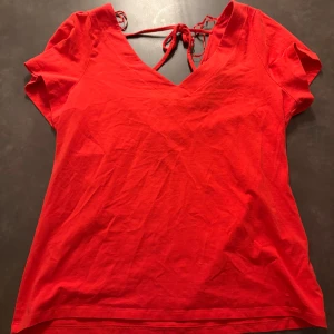 Röd topp med knyt i ryggen - Röd kortärmad topp med v-ringning och snygg knytning i ryggen. Tillverkad i mjuk bomull och har en enkel, stilren design som passar perfekt till jeans eller kjol. Stolek L men passar till en M! 