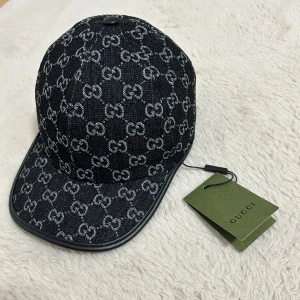 Gucci Keps - Snygg svart keps från Gucci med ikoniskt GG-mönster i silvergrått över hela kepsen. Kepsen har böjd skärm, justerbar rem bak i svart läder och detaljer i svart. Materialet är denim med läderdetaljer, perfekt för dig som gillar lyxiga accessoarer.
