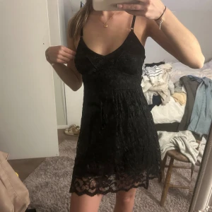 Svart spetsklänning med smala axelband - Supersöt svart kort klänning i spets med smala axelband och v-ringning. Klänningen har en figurnära passform upptill och lätt utsvängd kjol med fin spetskant nertill. Perfekt för dig som vill ha en stilren och feminin look.
