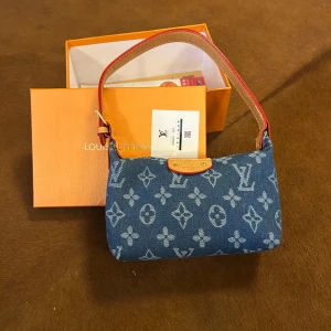 Louis Vuitton denim axelväska - Snygg axelväska från Louis Vuitton i blå denim med ikoniskt monogrammönster. Väskan har detaljer i ljust skinn, guldfärgad dragkedja och en kort axelrem med röda kanter. Perfekt storlek för det viktigaste och en riktigt trendig accessoar.