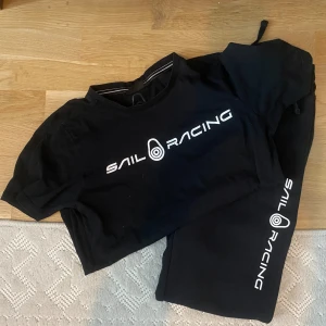 Svart Sail Racing set-  t-shirt + träningsbyxor  - Svart t-shirt från Sail Racing med vit logga tryckt över bröstet. Klassisk rund hals och korta ärmar, perfekt för dig som gillar sportig och clean stil. Materialet är mjuk bomull som känns skönt mot huden. Säljs som matchande set med träningsbyxor i bomull med dragsko och fickor.