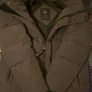 Svart pufferjacka från Canada Goose - Säljer en svart pufferjacka från Canada Goose med snyggt broderat märke på ärmen och avtagbar päls på huvan. Jackan har dragkedja, knappar och är långärmad. Perfekt för kalla vinterdagar och har en clean, mörk look.