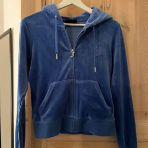 Blå velour hoodie Juicy Couture XS - Blå hoodie från Juicy Couture i mjuk velour med huva och snörning. Tröjan har dragkedja med JC-logga, fickor framtill och ribbade muddar. Perfekt för chill dagar och har en klassisk streetstil.