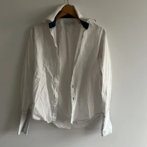 Vit skjorta med broderi från Rose Loco - Snygg vit skjorta från poco Loco i storlek M med mörkblå detaljer på kragen och tunna svarta sömmar. Skjortan har paisleybroderi längs knappraden, klassisk krage och långa ärmar med knappar vid manschetten. Perfekt för dig som gillar stilrena plagg med unika detaljer.