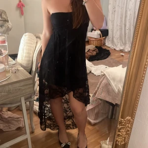 Svart spetsklänning från Club L - Säljer en svart långklänning från Club L med strapless design och transparent spets nedtill. Klänningen har en asymmetrisk form med kortare framdel och längre bakdel, vilket ger en cool och elegant look. Perfekt för dig som vill sticka ut med snygga detaljer.