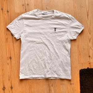 Vit t-shirt från Saint Laurent - Vit t-shirt från Saint Laurent med klassisk rund hals och svart YSL-logga broderad på bröstet. Enkel och stilren design som passar perfekt till jeans eller shorts. Tillverkad i mjuk bomull för en skön känsla hela dagen.