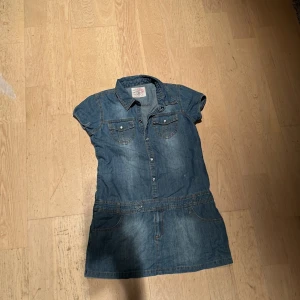 Jeansklänning Royal College Vintage Sports - Snygg jeansklänning med kort ärm, skjortkrage och knappar framtill. Två bröstfickor med lock och söta detaljer. Perfekt för dig som gillar en avslappnad men cool stil. Klassisk blå denim och praktiska fickor framtill.