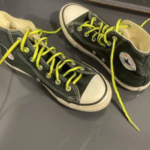 Converse All Star svarta sneakers - Svarta Converse All Star high-top sneakers med neongula snören och vit gummisula. Klassisk rund tå och tydliga vita sömmar. Märkets logga på sidan och All Star-tryck på hälen. Skorna är tillverkade i bomullstyg med platt sula.