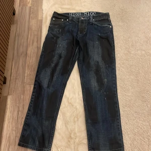 HIGH H100 jeans med L-detalj, blå - Unika blå jeans från HIGH H100 med coolt slitet och tvättat utseende. Snygga vita broderade detaljer på midjan och bakfickorna, samt klassisk femficksdesign. Raka ben och normal passform, tillverkade i robust denim. Perfekt för dig som gillar streetwear och vill sticka ut.