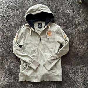 Grå hoodie från Vinson Polo Club - Grå hoodie med hel dragkedja från Vinson Polo Club. Broderad logga med brittisk flagga och guldfärgade detaljer på bröstet, samt broderad siffra och polospelare på ärmarna. Storlek S men liten i storleken så sitter mer som XS