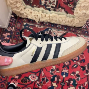 Adidas Samba sneakers strl 41 1/3 - Klassiska Adidas Samba sneakers i beige och svart med bruna gummisulor. Skorna har svarta ränder på sidorna, svart häl och snörning. Tillverkade i skinn och syntet, perfekt för dig som gillar retrostil och streetwear.