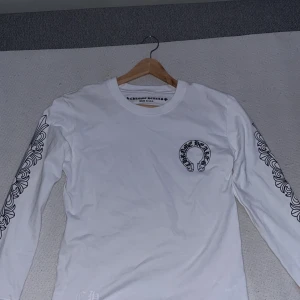 Vit långärmad tröja Chrome Hearts - Vit långärmad tröja från Chrome Hearts med svart tryck på bröstet och detaljerade grafiska mönster längs båda ärmarna. Rund hals och tillverkad i mjuk bomull. Perfekt för dig som gillar streetwear och snygga detaljer.