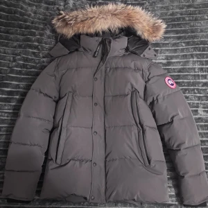 Canada Goose med äkta Päls - Säljer en mörkgrå dunjacka från Canada Goose med äkta päls på huvan. Jackan har klassisk puffer-design, lång ärm och logotyp på ärmen. Fodrad insida och praktiska fickor framtill. Perfekt för kalla vinterdagar och riktigt snygg streetstil.