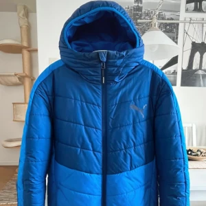 Blå pufferjacka från Puma med huva - Snygg blå pufferjacka från Puma med huva och hel dragkedja framtill. Jackan har quiltad design och logga på bröstet. Perfekt för kalla dagar och har en sportig vibe. Färgen är klarblå med mörkare blå detaljer upptill. Passformen är normal och jackan är långärmad.