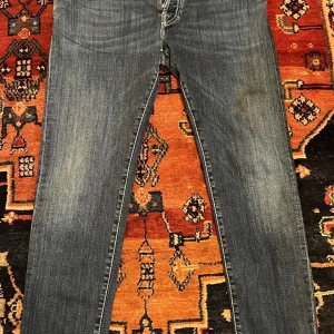 Jacob Cohën blå jeans med röd detalj - Snygga blå jeans från Jacob Cohën med klassisk femficksdesign och kontrastsömmar i rött. Jeansen har en röd patch med logga bak och unika knappar framtill. Materialet är slitstarkt denim och passformen är rak med normal midja.