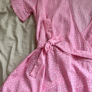 Rosa prickig omlottklänning Vero Moda - Superfin rosa omlottklänning från Vero Moda med vita prickar. Klänningen har volangkant nertill, knytband i midjan och korta ärmar. Perfekt för dig som gillar en feminin och lekfull stil.