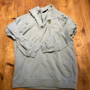 Ljusgrå tröja från Lyle & Scott - Säljer en ljusgrå långärmad tröja från Lyle & Scott med klassisk gul logga på bröstet. Tröjan har krage med halv dragkedja och är tillverkad i mjuk bomull. Perfekt för lager-på-lager och chill vardagsstil.