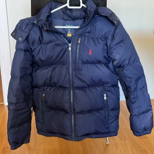 Mörkblå pufferjacka från Polo Ralph Lauren - Snygg mörkblå pufferjacka från Polo Ralph Lauren med röd logga på bröstet. Jackan har dragkedja framtill, två sidofickor med dragkedja och elastiska muddar vid ärmslut. Perfekt för kalla dagar och har en klassisk, sportig look.