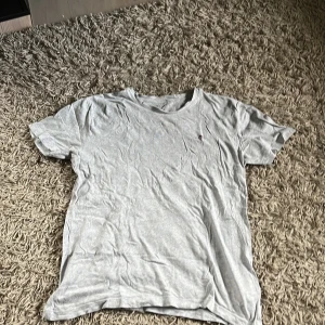 Ljusgrå t-shirt från GANT - Ljusgrå t-shirt från GANT med klassisk passform och rund hals. T-shirten har ett litet tryckt GANT-logo med sköld på bröstet. Tillverkad i mjuk bomull, perfekt för en clean och enkel stil.