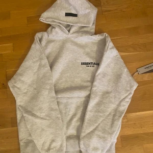 Grå hoodie Essentials Fear of God - Snygg grå hoodie från Essentials Fear of God med svart logga på bröstet och ryggen. Tröjan har huva, ribbade muddar och en klassisk loose passform. Perfekt för dig som gillar streetwear och vill ha en stilren look. S men passar M typ