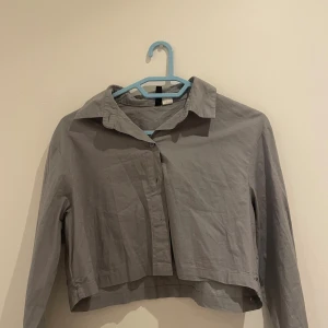 Grå croppad skjorta med knappar - Snygg grå croppad skjorta med klassisk krage och knappar framtill. Skjortan har lång ärm och är tillverkad i ett lätt bomullstyg som känns skönt mot huden. Perfekt att styla med höga jeans för en trendig look.