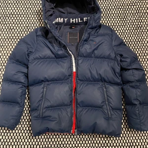 Mörkblå dunjacka Tommy Hilfiger - Mörkbå dunjacka från Tommy Hilfiger. Jackan har dragkedja med röd detalj, två sidofickor med dragkedja. Fodrad med dun och fjäder. Perfekt för kalla dagar. 
