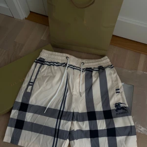 Burberry badshorts - Helt oanvända snygga beiga badshorts från burberry. Aldrig använda med etiketten kvar. Säljes då dem är en present som inte kommit till användning. Storleken är S men ganska stor och passar därav en M. Sitter perfekt på mig som är 190cm och 85kg.