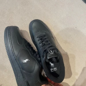 Svarta sneakers från Polo Ralph Lauren - Snygga helsvarta sneakers från Polo Ralph Lauren med klassisk logga på sidan och tungan. Skorna har snörning, rund tå och är tillverkade i skinn med detaljer i syntet. Perfekta för dig som gillar stilrena och sportiga skor.