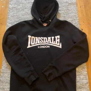 Svart Lonsdale hoodie med tryck - Svart hoodie från Lonsdale med stort vitt Lonsdale London-tryck på bröstet. Klassisk huva med snörning och ribbade muddar vid ärmslut och nederkant. Perfekt för dig som gillar streetwear och sportig stil.