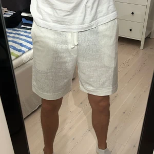 Vita shorts med snörning i linne-look - Säljer ett par fräscha vita shorts med relaxed fit och snörning i midjan. De har en lätt textur som påminner om linne, sidofickor och en bakficka.