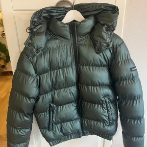 Grön pufferjacka från Goodfornothing - Snygg grön pufferjacka från Goodfornothing med huva och dragkedja framtill. Jackan har en cool, lite glansig yta och två fickor med dragkedja. Perfekt för dig som vill ha en varm och trendig jacka med loose passform och lång ärm. Nytvättat och inga tecken på användning