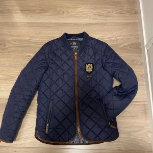Mörkblå quiltad jacka från Morris - Snygg mörkblå quiltad jacka från Morris med bruna detaljer och emblem på bröstet. Jackan har dragkedja framtill, två fickor med dragkedja och ribbad krage. Insidan har ett paisleymönstrat foder som ger extra stilpoäng. Perfekt för dig som gillar klassisk look.