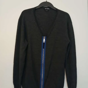 Svart kofta från Raf Simons med dragkedja - Säljer en svart kofta från Raf Simons med unik blå detalj längs dragkedjan framtill. Koftan har v-ringning, lång ärm och är tillverkad i ett tunt, stickat material. Perfekt för dig som gillar stilrena plagg med en twist.