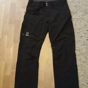 Svarta friluftsbyxor från Haglöfs - Svarta friluftsbyxor från Haglöfs med rak passform och diskret logga på vänster lår. Byxorna har knappstängning och dragkedja fram samt praktiska fickor med dragkedja. Perfekta för utomhusaktiviteter och äventyr.