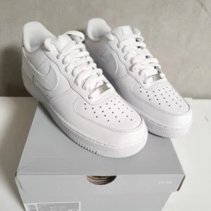 Nike Air Force 1 helvita sneakers - Nike Air Force 1 sneakers i helvitt med klassisk låg siluett. Skorna har snörning, perforerad tå och platt sula med tydlig struktur undertill. Tillverkade i skinn med ikonisk Swoosh på sidan. Perfekt för dig som gillar cleana och tidlösa sneakers.