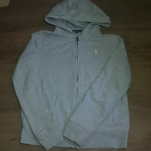 Grå hoodie från Polo Ralph Lauren - Snygg grå hoodie från Polo Ralph Lauren med dragkedja och klassisk broderad logga på bröstet. Tröjan har huva, fickor framtill och är tillverkad i mjuk bomull. Perfekt för en chill och stilren look.