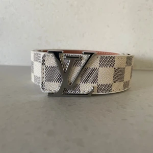 L och V bälte 105cm  - L och V bälte med ikoniskt rutigt mönster i beige och grått. Bältet har en stor L och V logga i silverfärgad metall som spänne. Perfekt accessoar för dig som vill sticka ut med en lyxig detalj.
