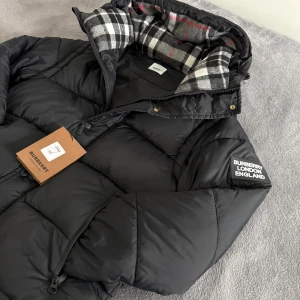 Burberry Jacka  - Svart Dunjacka från Burberry med avtagbara ärmar, vilket gör den till en väst också. Jackan har dragkedja, huva med rutigt foder i svart, vitt och grått samt en tydlig Burberry London England patch på ärmen. Praktiska fickor med dragkedja och quiltad design.