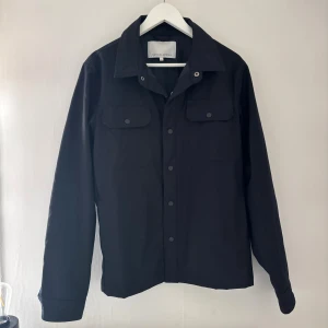 Svart overshirt från Casual friday - Stilren svart overshirt från Casual Lover med klassisk krage och tryckknappar framtill. Jackan har två bröstfickor med lock och långa ärmar. Perfekt för lager-på-lager och enkel att matcha med olika outfits.