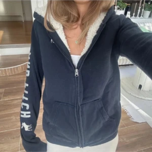 Mörkblå teddyjacka Abercrombie - Mysig mörkblå teddyjacka från Abercrombie & Fitch med fluffigt vitt foder och huva. Jackan har dragkedja framtill, två fickor och stora vita logotryck längs båda ärmarna. Perfekt för kalla dagar och chill stil.