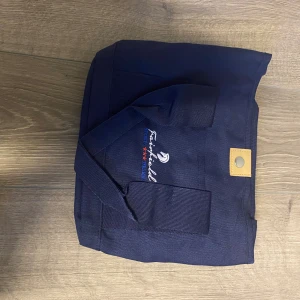 Mörkblå Fairfield ryktväska - Mörkblå shoppingväska från Fairfield med broderad logga och två röda stjärnor. Väskan har kraftiga handtag och en tryckknapp i läderimitation för stängning. Perfekt för att ha som ryktväska.