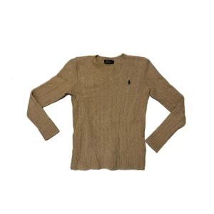 Beige kabelstickad tröja Polo Ralph Lauren - Snygg beige kabelstickad tröja från Polo Ralph Lauren med klassisk rund hals och broderad marinblå logga på bröstet. Tröjan har långärmad passform och ribbade muddar vid ärmslut och nederkant. Perfekt för dig som gillar stilren och tidlös design.