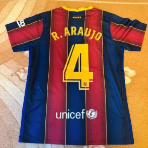 FC Barcelona R. Araujo matchtröja Nike - Säljer en officiell FC Barcelona fotbollströja från Nike med R. Araujo och nummer 4 på ryggen. Tröjan har klassiska blå och röda vertikala ränder med gula detaljer, klubbmärke på bröstet och sponsorer. Tillverkad i lätt och ventilerande material.