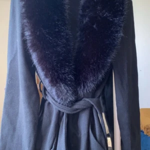 Svart kappa med päls och bälte - Snygg svart kappa med bred krage i mjuk pälsimitation och knytbälte i midjan. Perfekt för dig som vill ha en elegant och varm look under kyliga dagar. Långärmad och klassisk design som ger en lyxig känsla.