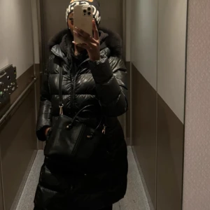 Svart lång colmar med päls  - Säljer en svart, lång pufferjacka med glansig yta och stor huva med pälsdetalj. Jackan har dragkedja framtill och är perfekt för kalla dagar. Den har en oversized passform och långa ärmar, vilket gör den extra mysig och trendig.