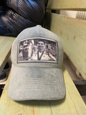 Grå meshkeps från Berretto Ronaldo keps  - Snygg grå keps från Berretto med mjuk mockaliknande front och mesh på sidorna och bak. Framtill finns ett svartvitt tryck med sportmotiv och broderad logga. Justerbar baktill för perfekt passform. Perfekt för dig som gillar sportig stil.