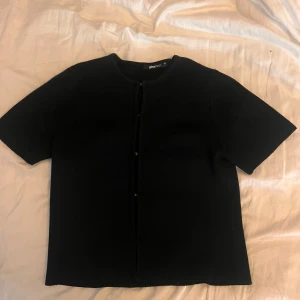 Svart kortärmad kofta från Gina Tricot - Svart kofta från Gina Tricot med korta ärmar och rund hals. Koftan har diskreta hyskor framtill istället för vanliga knappar, vilket ger en clean look. Materialet känns mjukt och har en lätt stickad känsla, perfekt för lager-på-lager-stil.