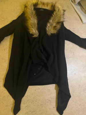 Svart teddyjacka med fuskpäls från SHEIN - Svart teddyjacka från SHEIN med stor, fluffig fuskpäls i bruna toner runt kragen. Jackan har lång ärm och en loose passform, perfekt för att hålla sig varm och snygg under kyliga dagar. Materialet är mjukt och mysigt, och kragen ger en extra cool touch.